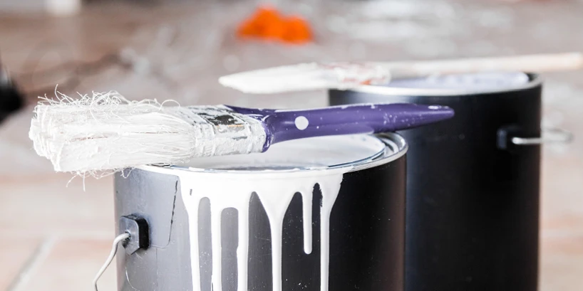 Photo of two black paint cans with a used purple-handled paint brush resting on top, white paint is spilling out of the front paint can