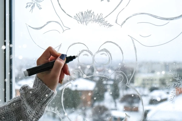 Woman using white paint marker to paint holiday deer on window with snow falling outside.