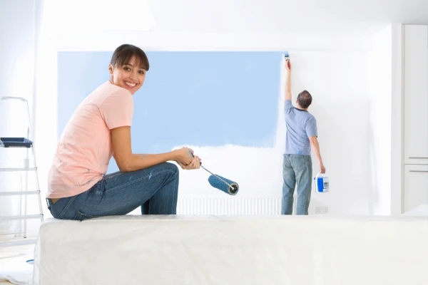 Smiling woman holding a paint-roller in her hand while her husband is applying blue paint to the living room wall using a paint brush